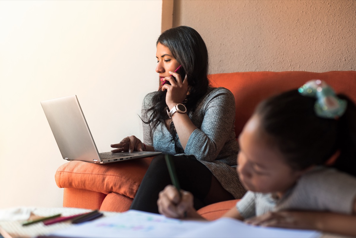 woman on cell phone looking at laptop while child does homework in foreground