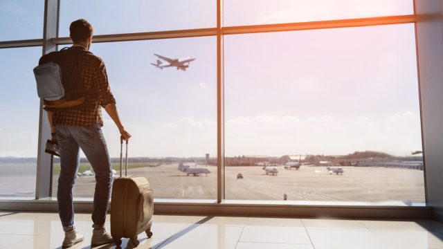 Calm male tourist is standing in airport and looking at aircraft flight through window. He is holding tickets and suitcase. Sunset