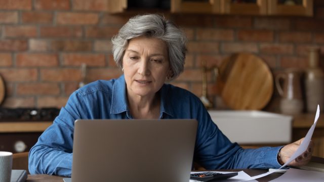 Smiling mature woman using laptop, online baking service, checking financial documents at home, senior grey haired female sitting at table with domestic bills and calculator, accounting
