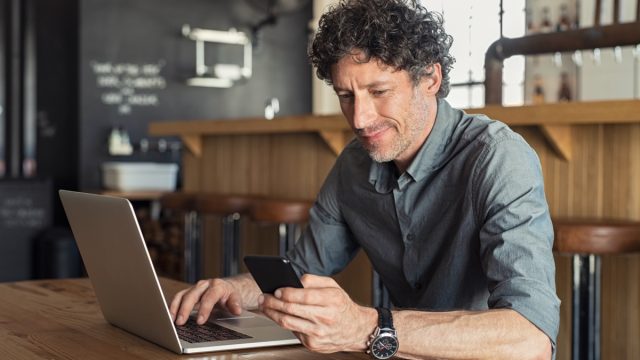 Happy mature business man sitting at cafeteria with laptop and smartphone. Businessman texting on smart phone while sitting in a pub restaurant. Portrait of senior formal man working and checking email on computer.