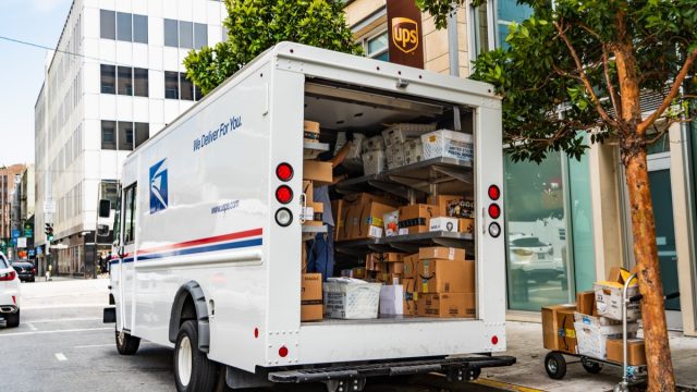 USPS delivery van stopped in front of a UPS location, unloading Amazon packages