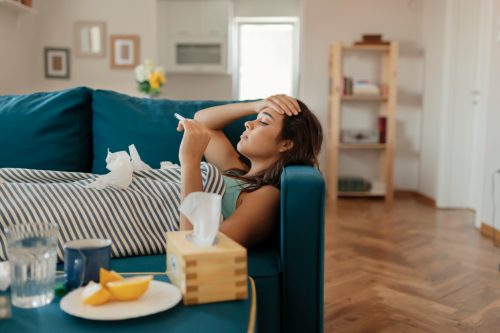 A young woman lying sick on the couch and checking her temperature