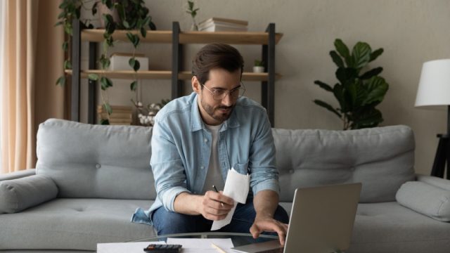 man holding a piece of paper with papers next to him while using his laptop on the couch to do taxes
