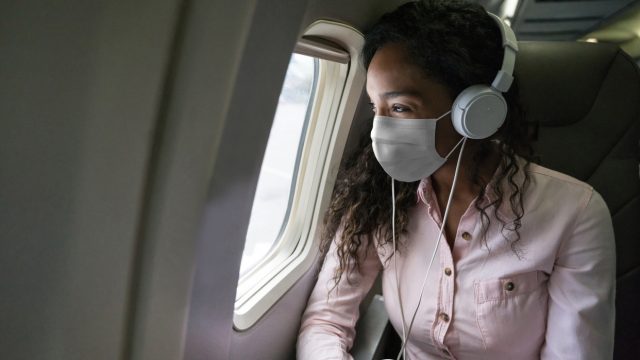 A young woman wearing a mask and headphones looking out the window of an airplane