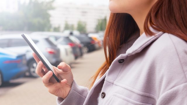 woman holding mobile phone in hand standing near cars parking lot. Searching information and news. Social media concept.