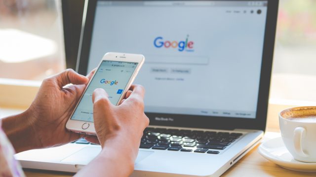 A close up of someone's hands using a phone and a laptop to access Google