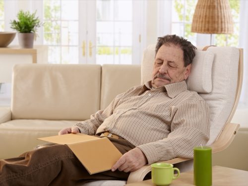Older man napping on a chair in his living room with a book on his lap