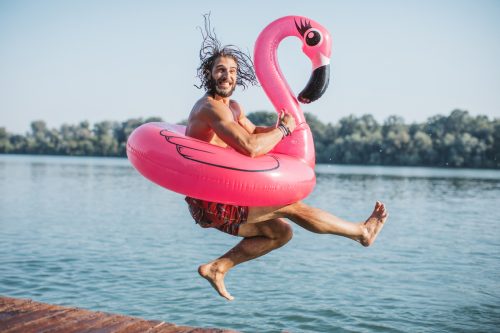 Young man jumping in river from deck. He is jumping with pink flamingo swimming float.