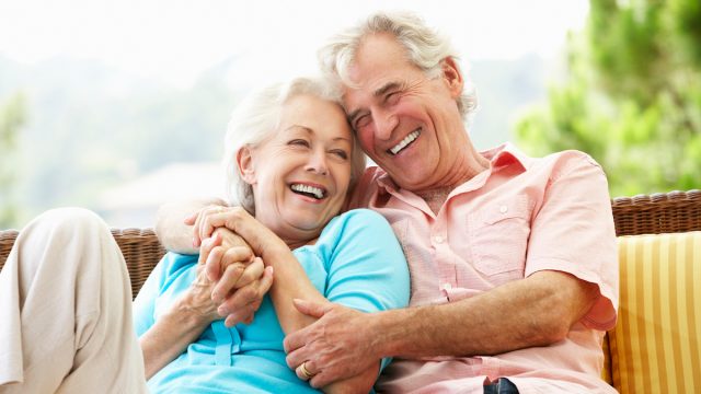 An older couple, both with white hair, sitting on a bench, hugging and laughing.