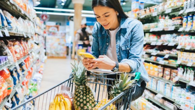 Happy young woman shopping for groceries in supermarket standing near her trolley using smartphone