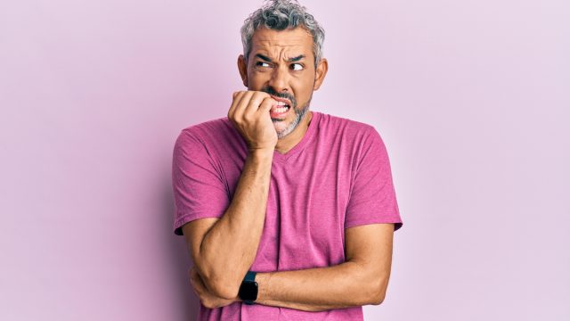 A middle-aged man with gray hair and a purple t-shirt nervously biting his nails looking insecure against a light purple background