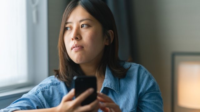 A young woman sitting on a sofa by the window and using a smart phone st home.