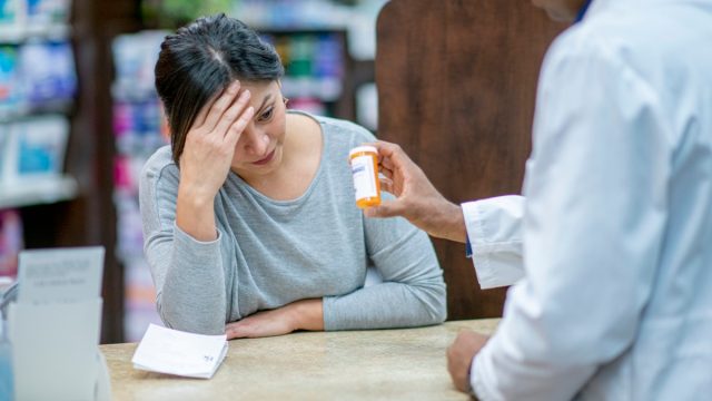 A stressed customer with her head in her hand leans on the counter and discusses medication with the pharmacist.