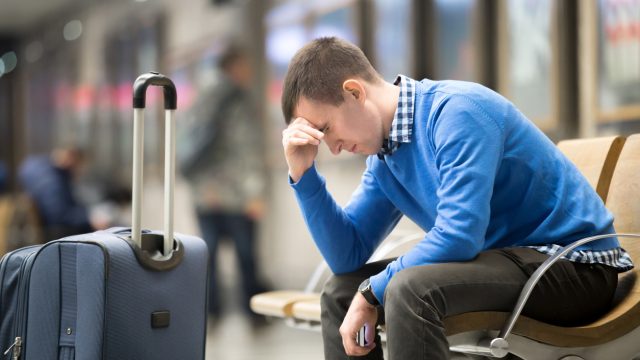 A man sitting in an airport with his luggage holding his head looking distressed