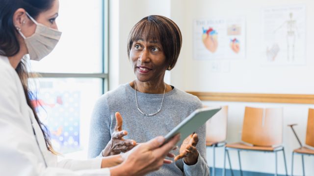An attentive doctor listens as a female patients asks questions about recent medical test results. The doctor is holding a digital tablet which contains the test results. The doctor is wearing a protective face mask.
