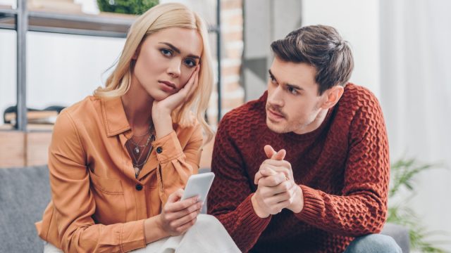 Husband looking at unhappy young woman holding smartphone and looking at camera at home, relationship problem concept