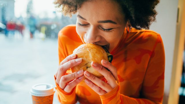 A woman sitting in a cafe eating a bagel while wearing a bright orange shirt.