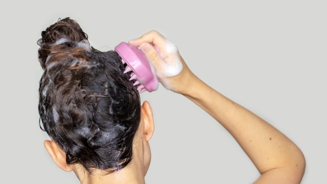 Close up of the back of a woman's head in the shower with shampoo in her hair and using a scalp massager.