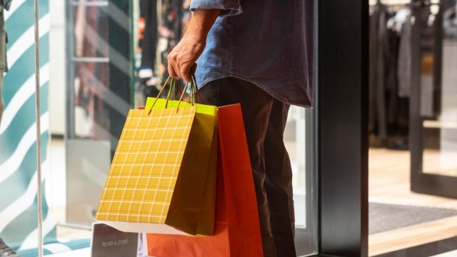 Senior man in a denim shirt holding many shopping bags looks at the illuminated windows and enjoys evening shopping.