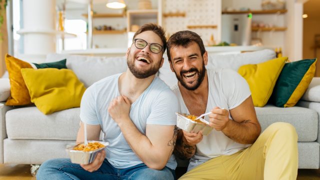 A male couple eating pasta out of takeout containers while sitting on their living room floor and laughing.