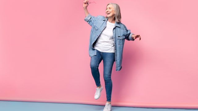 An older, gray-haired woman wearing skinny jeans and a denim shirt dancing happily on a pink background.