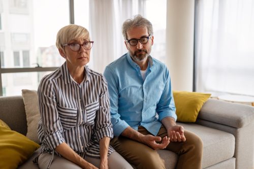 Mature man and woman sitting on a couch in their apartment looking forlorn
