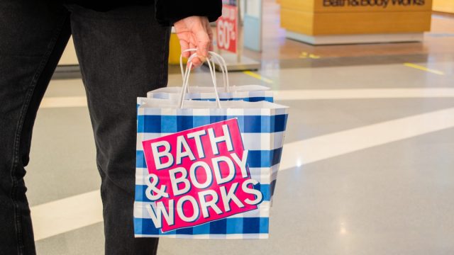 A female customer holds in her hand paper bags branded “Bath and body works” in a blue and white check and a pink logo on the background of the entrance to the store