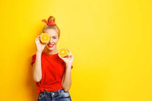 A young woman in a red t-shirt with her blonde hair tied up in a red bow, holding two orange slices against a yellow background.