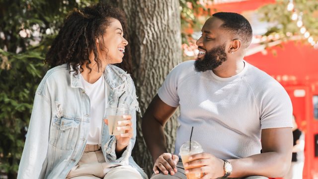 A couple on a date is sitting outside in front of a tree smiling while drinking lemonade.