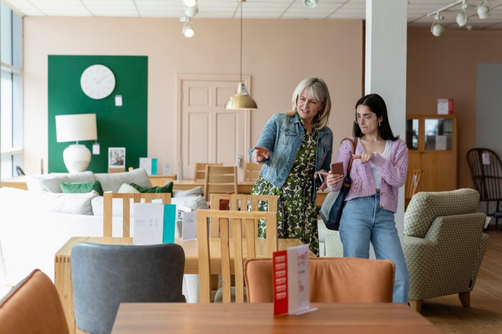A mature woman and her daughter looking around a furniture store together for new furniture for home. They are looking for an item that the daughter has up on her phone, that she has seen online.