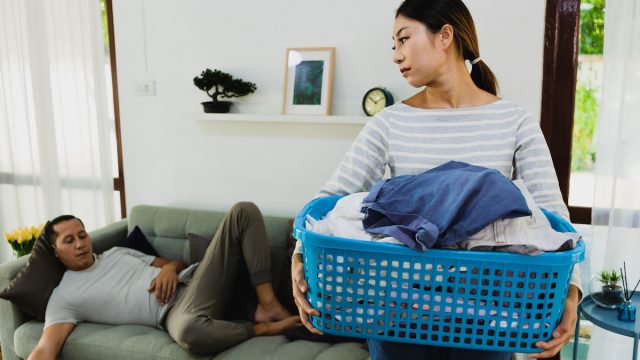 An annoyed-looking wife carries the laundry basket while her husband sleeps on the couch.