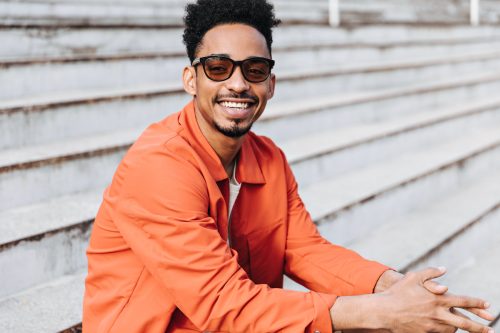 A cheerful man wearing an orange button-down shirt smiling and wearing sunglasses while sitting on outdoor steps