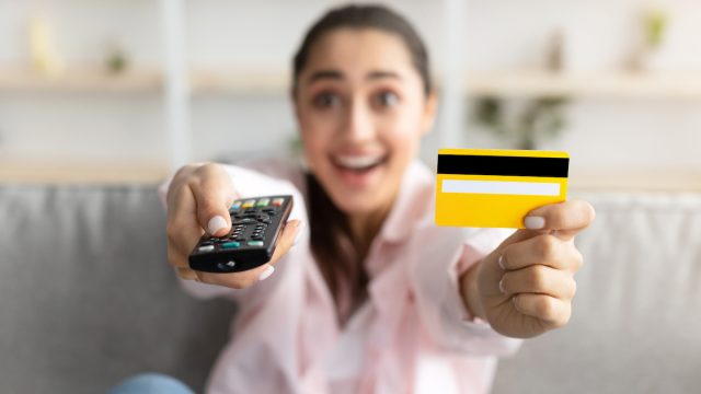 Closeup of excited millennial woman sitting on couch in living room, using tv remote and holding yellow credit card