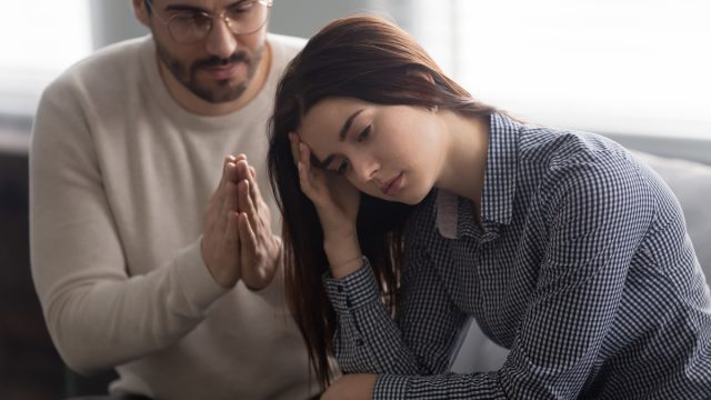 Husband sitting on couch with his wife and pleading for forgiveness with hands in prayer.