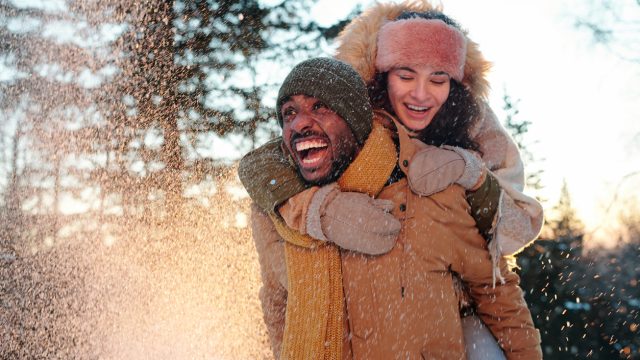 Cheerful multiracial couple in winterwear laughing while girl embracing her boyfriend during snowfall