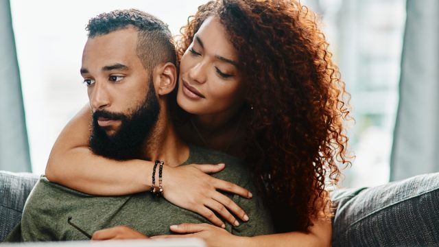 Shot of a young woman hugging her husband while he uses a laptop on the sofa at home