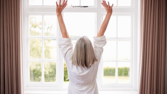 Rear view of a woman with gray hair wearing a white bathrobe stretching in the morning in front of a window