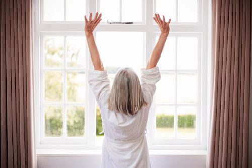 Rear view of a woman with gray hair wearing a white bathrobe stretching in the morning in front of a window