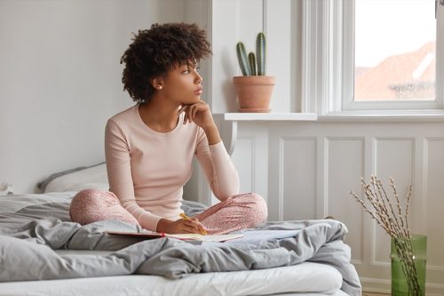 Woman manifesting, writing in her journal, sitting on her bed, looking out her window.