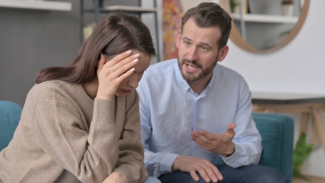 A couple on the couch arguing; the man is talking and the woman has her head in her hands.