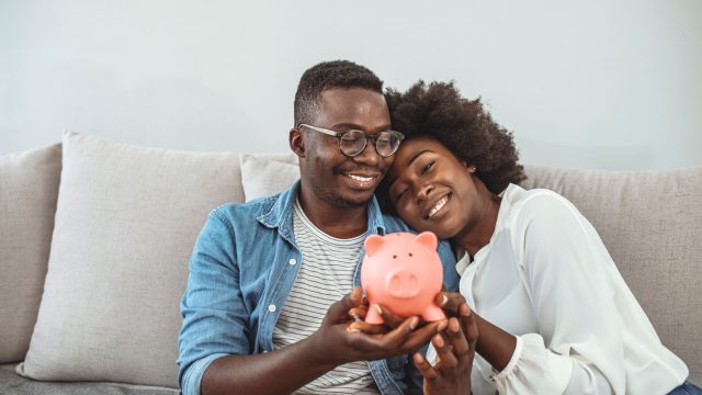 A couple sitting on the couch while smiling and holding a piggy bank
