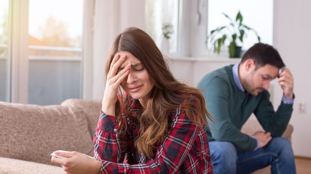 A young couple facing away from each other on the couch; the man has his head in his hands and the woman is crying.