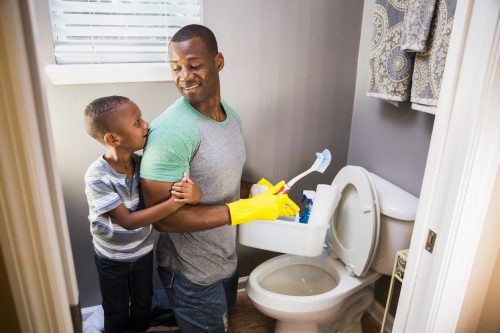 A young boy hugging the arm of a man cleaning a toilet while wearing yellow gloves and holding cleaning supplies