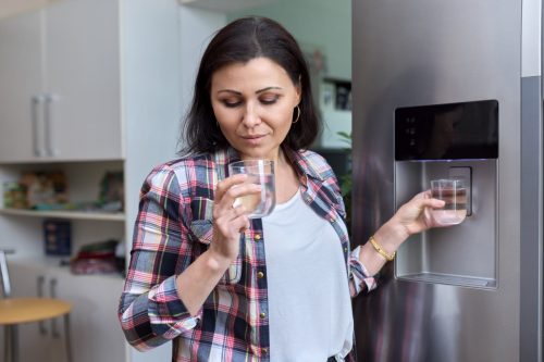 Water dispenser, woman taking cold water into glass from home refrigerator