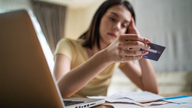 A young woman holding a credit card at her desk and looking at it with a sad expression