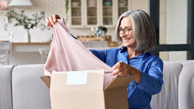 A woman with gray hair takes a shirt out of a box while sitting on her couch