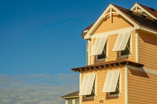 Close up of the top of a yellow house with storm shutters against a blue sky