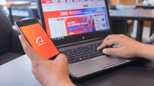 A person holds a phone with the Alibaba app open while shopping at Alibaba on their laptop.