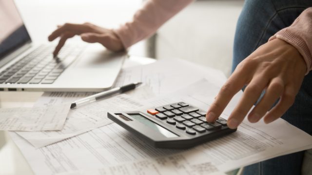 A closeup of someone using a calculator while inputing information onto a laptop