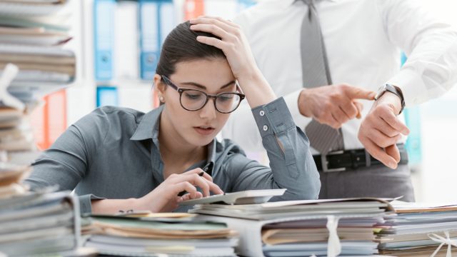 Demanding boss pointing to his watch and asking his employee to hurry up while she sits behind a stack of folders and papers